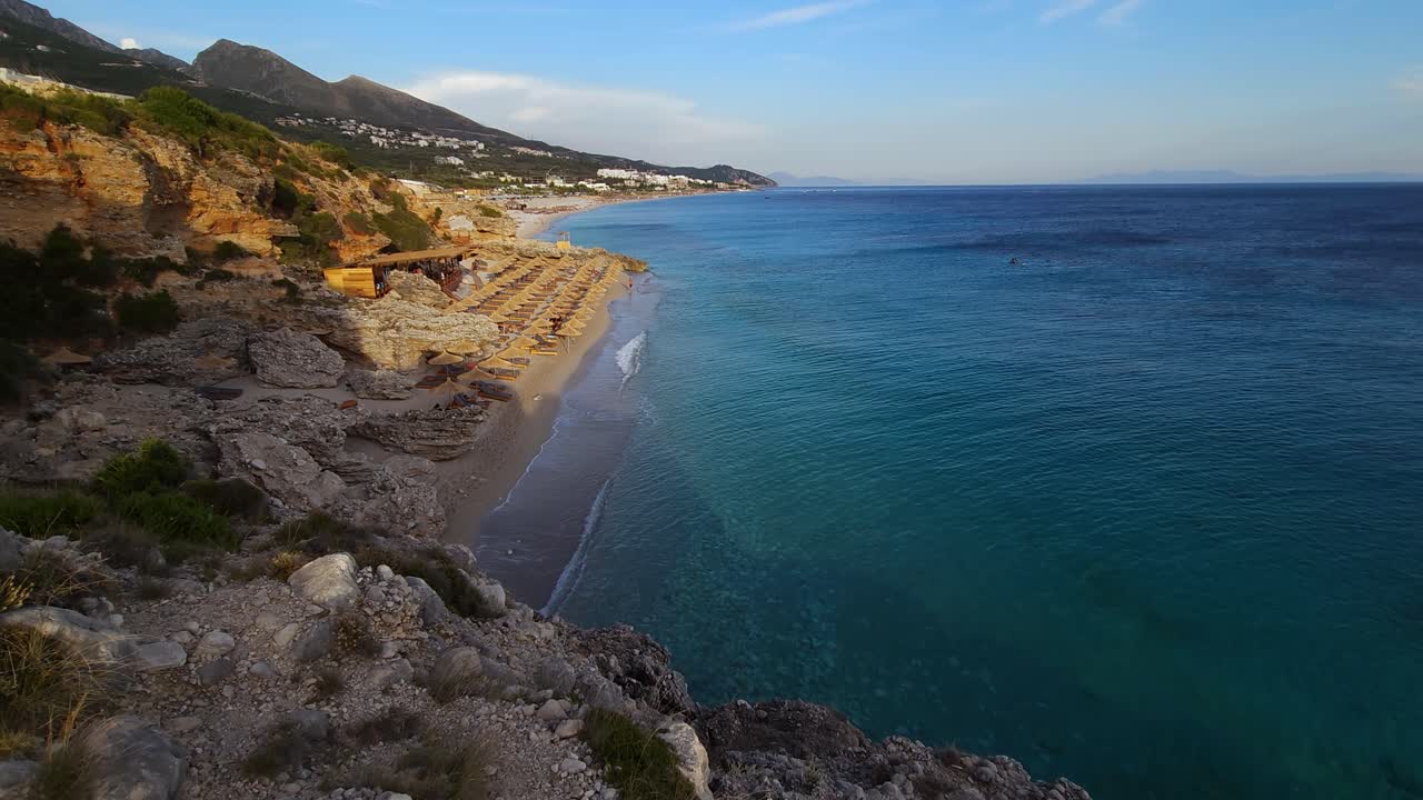 Arnavutluk'ta İyon Denizi kıyısında, hasır şemsiyeli güzel bir plajı aydınlatan güneş ışığı ve derin mavi denizin üzerindeki tepelerde yer alan turist tatil köyleri