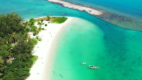 Anchored-boats-on-calm-clear-water-of-turquoise-lagoon-near-cape-of-tropical-island-with-white-sandy-beach-and-palm-trees,-Thailand