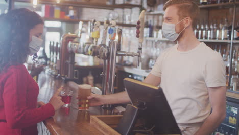 female customer wearing mask in bar making contactless payment for drinks during health pandemic