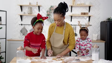 Retrato-De-Mamá-Sonriente-En-Delantal-Con-Niños-Pequeños-Parados-En-La-Mesa-En-La-Cocina-Decorada-De-Navidad-Y-Divirtiéndose-Mientras-Hacen-Galletas-De-Navidad