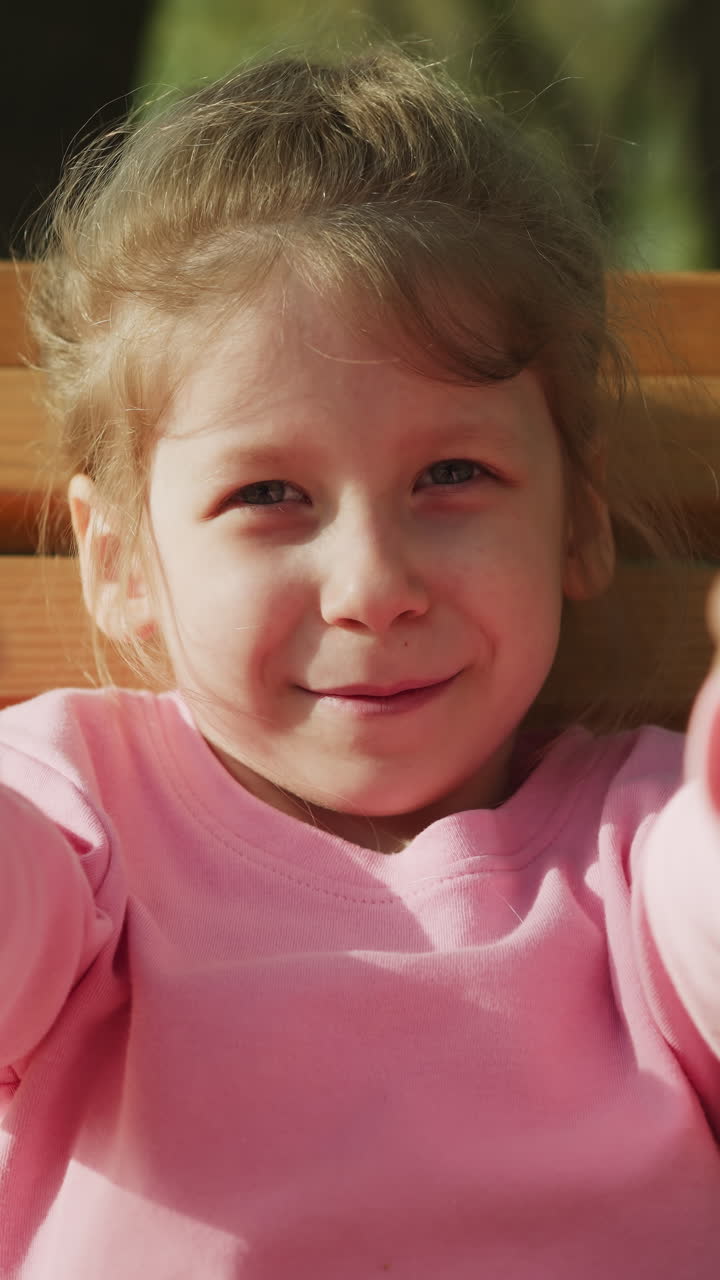 Smiling preschooler girl shows thumbs-up on wooden bench in spring park closeup. Everything will be OK gesture. Adorable blonde child in city garden