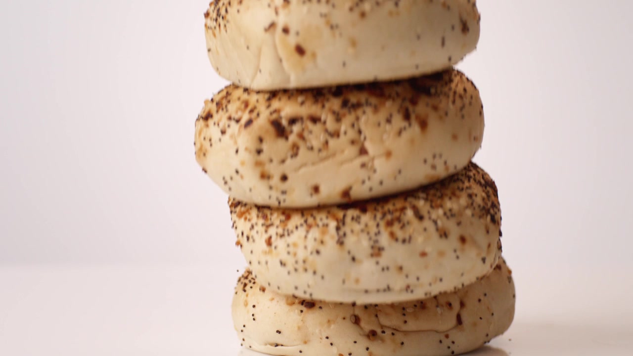 Bagel salati con semi di ogni tipo impilati che rimbalzano e rotolano su sfondo bianco in uno scatto macro da studio al rallentatore. Branding di gastronomia newyorkese, pubblicità di sandwich per la colazione, contenuti per negozi di bagel