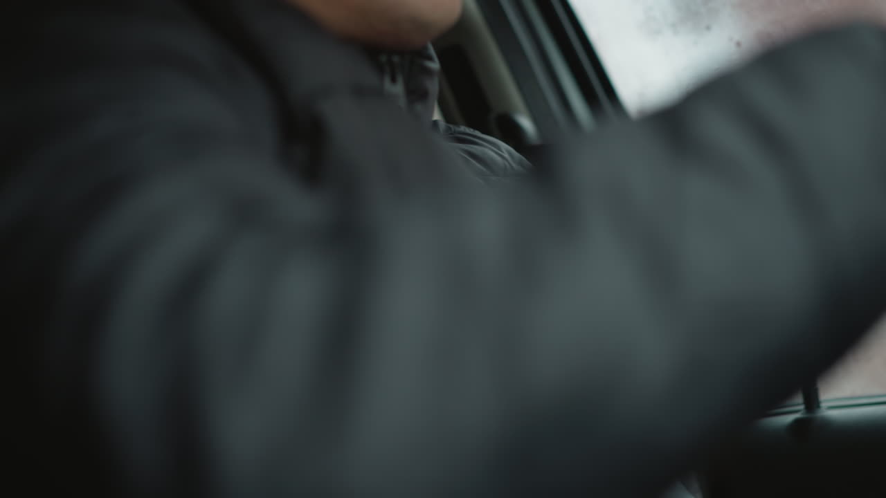 Close up of car buff wearing black winter coat pulling seat belt across torso while seated in vehicle cabin beside fogged window emphasizing safety ritual and hand gesture during cold commute