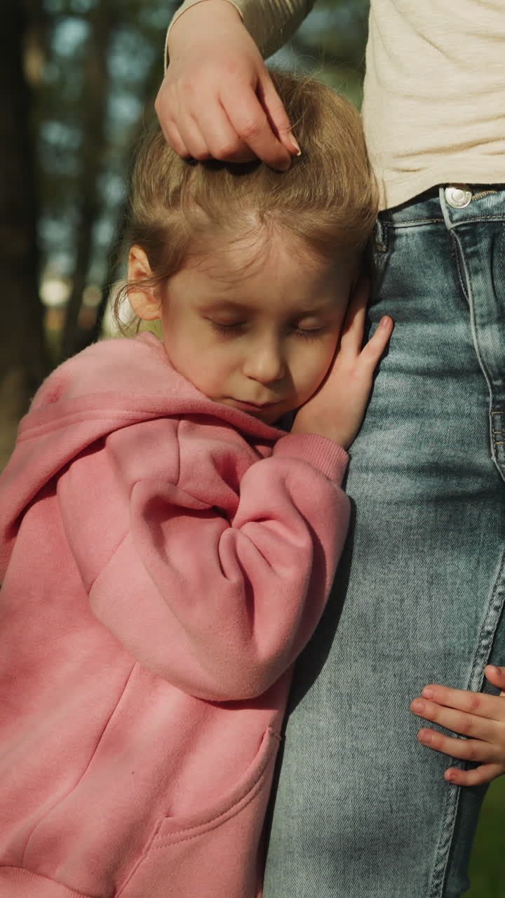 Sleepy little girl leans on mother thigh standing in spring park closeup slow motion. Woman and preschooler daughter after long walk in garden