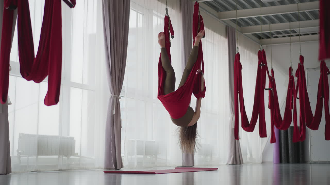 Female yoga instructor gently swings in red aerial silk hammock by floor to ceiling window, legs suspended mid air with slight sway left right, bright studio setting calm balance focus serenity