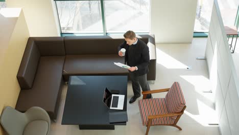 a professional stands in a modern office space holding a document and a coffee cup, analyzing colorful charts, with a laptop on the table and sunlight streaming through large windows - top down view