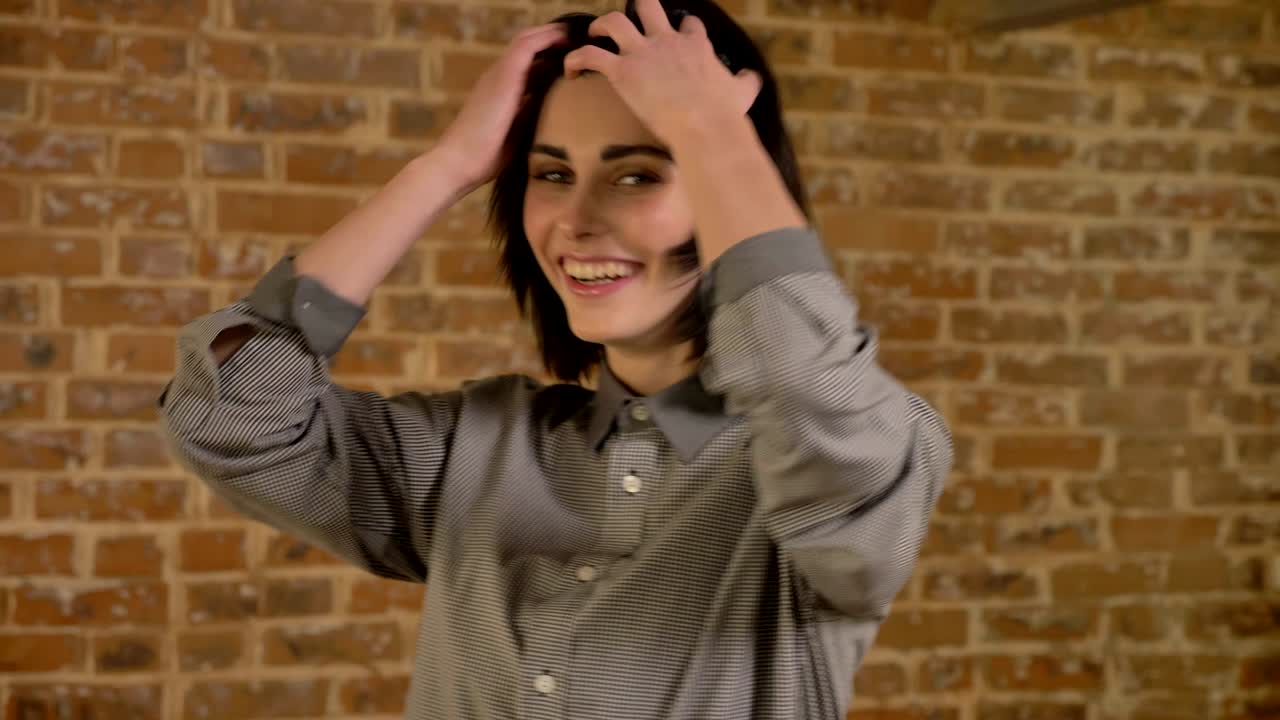 Young beautiful woman with short brown hair looking in camera and twisting around, jumping and laughing, happy, brick wall background