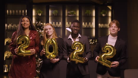 group of friends wearing elegant clothes holding balloons with the numbers 2022 while dancing in new year's party 2