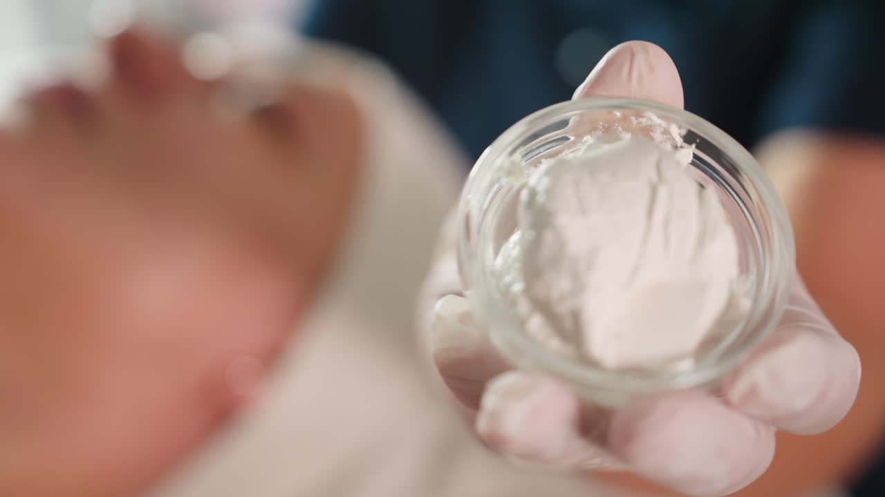 Gloved professional hand holding transparent silicone cup filled with creamy white facial mask, soft focus spa background, detailed closeup of treatment product in clinical setting