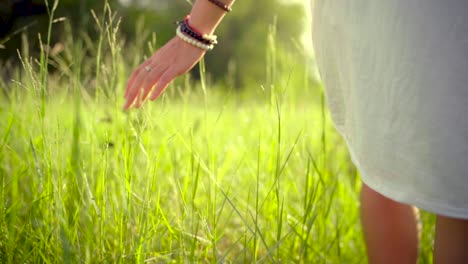 young woman in white dress walking on meadow and gently touching grass with morning dew at sunrise