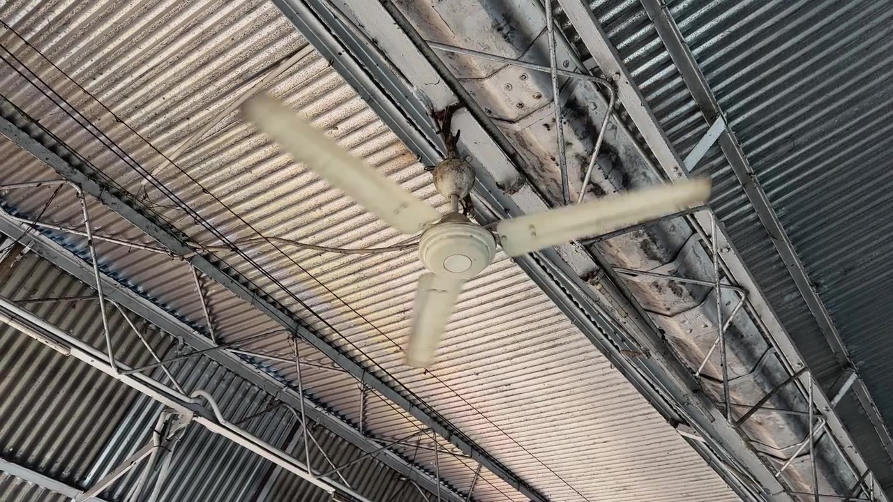 Close up of a rotating industrial ceiling fan mounted beneath a corrugated metal roof in a public transport facility