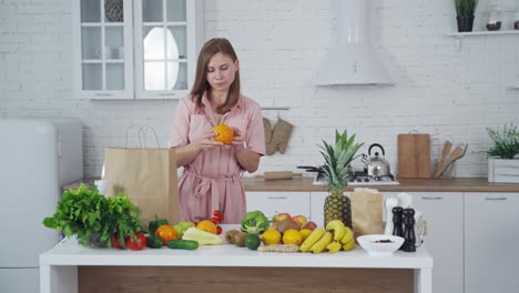 pretty woman taking out fresh fruit from paper shopping bag at home. young female holds an orange in the light kitchen. healthy fruit and vegetables on the table.