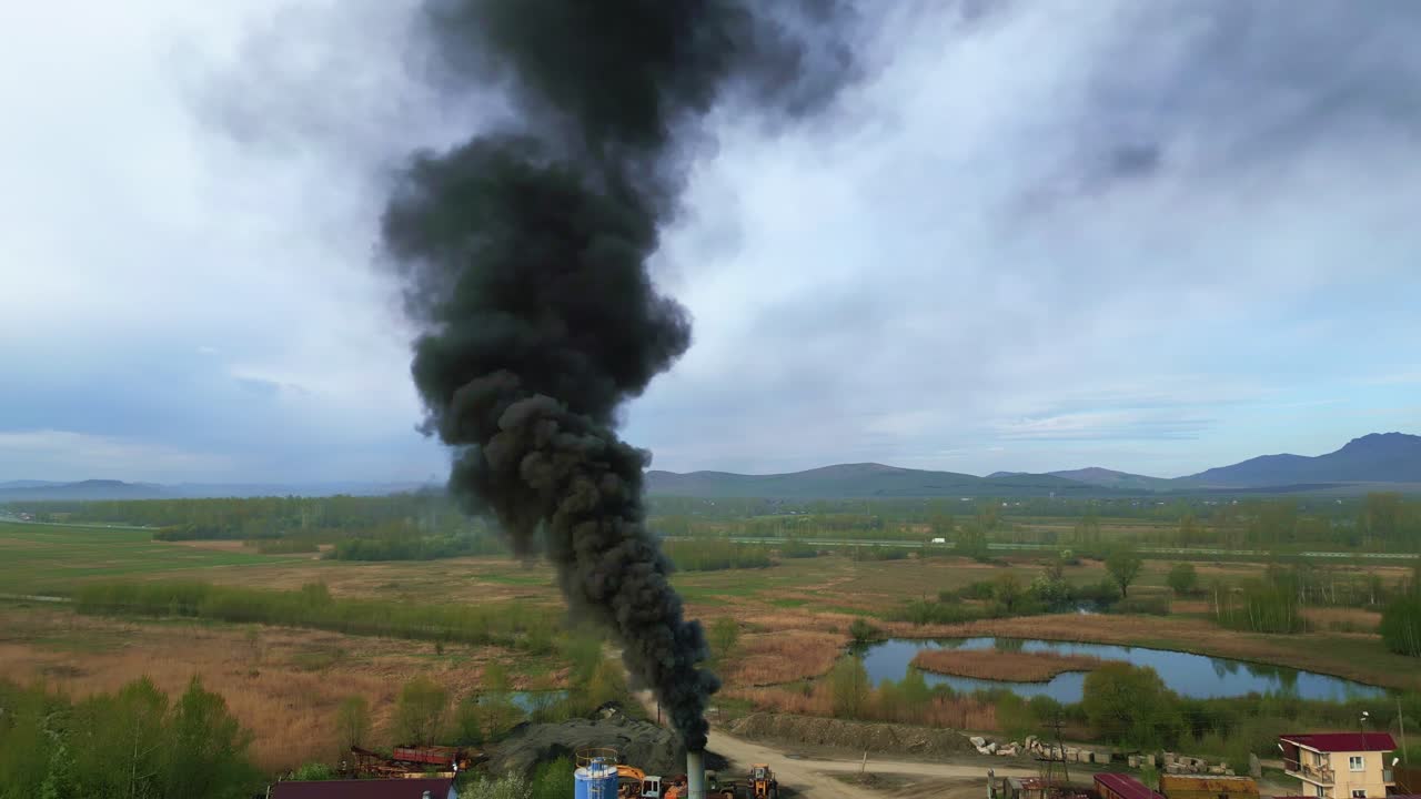 Fumo nero si innalza da un sito industriale distante. Rigogliose colline verdi circondano la scena. Un lago calmo riflette il cielo nuvoloso. Nessuna persona è visibile nel filmato.