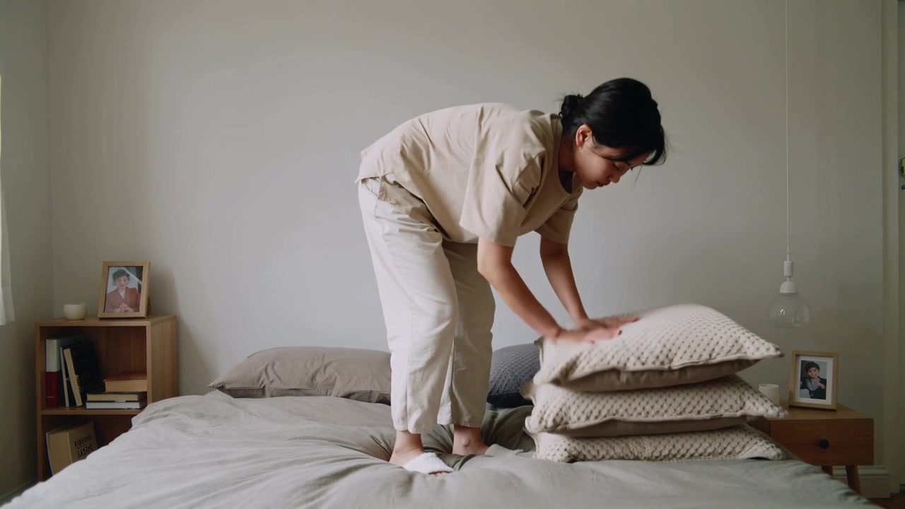 Memasuki kamar tidur, wanita Asia berdiri di tempat tidur menata bantal dekat nakas untuk merapikan tempat tidur.