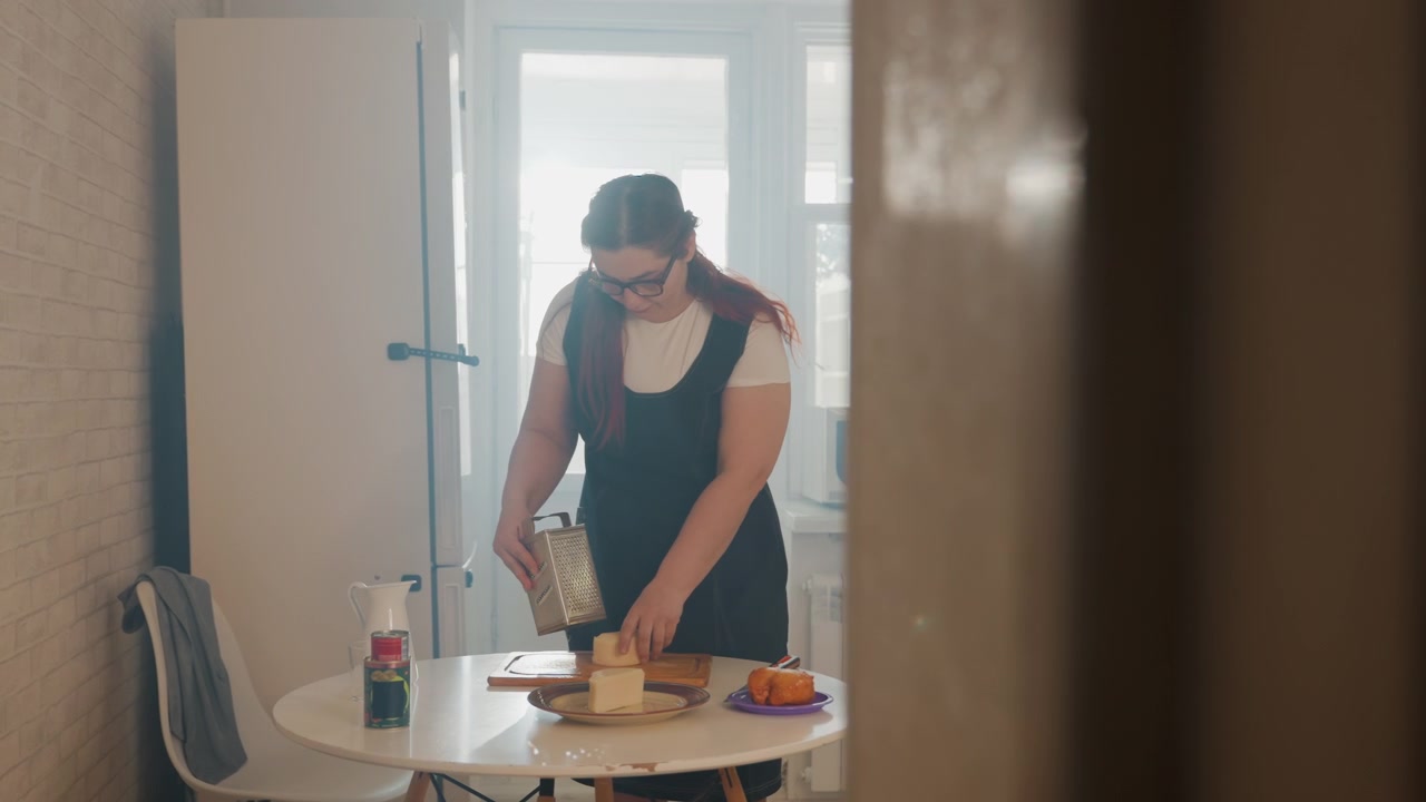 Donna in piedi a un piccolo tavolo rotondo da cucina prepara un pasto, affettando verdure su un tagliere, versando liquido in un misurino, con postura concentrata, cucina domestica ordinata e luce soffusa dalla finestra.