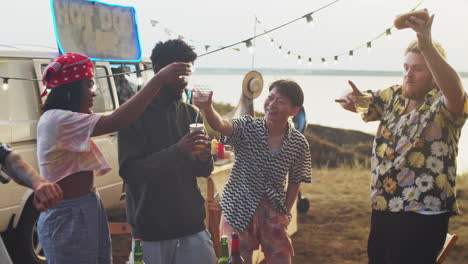 happy multiethnic friends dancing with drinks at summer festival