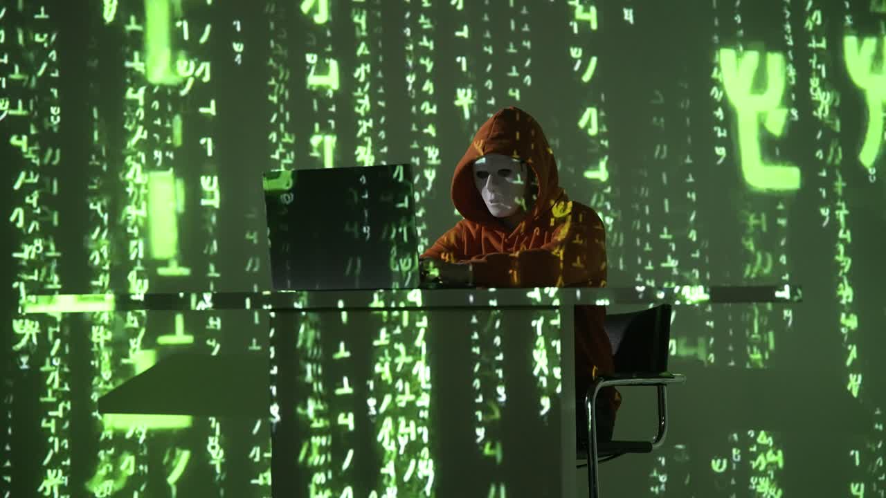 A masked hacker in an orange hoodie sits at a desk with a laptop, surrounded by projected green code. The scene illustrates themes of cybercrime, hacking, and digital surveillance