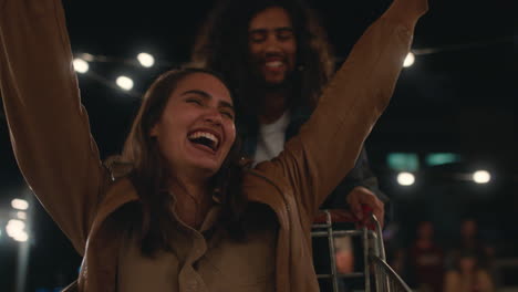 Hermosa-Joven-Divirtiéndose-Sentada-En-Un-Carrito-De-Compras,-Grupo-De-Amigos-Disfrutando-De-Una-Loca-Fiesta-En-La-Azotea-Por-La-Noche-Celebrando-Juguetonamente-El-Fin-De-Semana