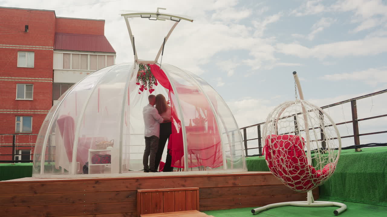 romantic rooftop dome date couple outdoors intimate moment inside transparent bubble dome decorated with red roses and string lights, city building backdrop, cozy seating, champagne toast, sunset sky