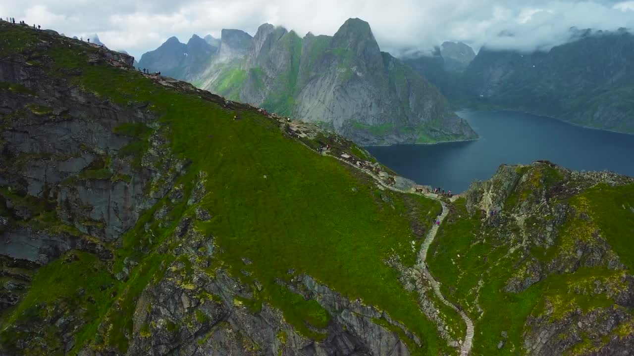 Aerial drone footage flying closer to tall steep rocky cliffs and mountains in Lofoten Norway, large blue river or water lake is in betwen mountains. Mountain has hiking trails and small people hiking
