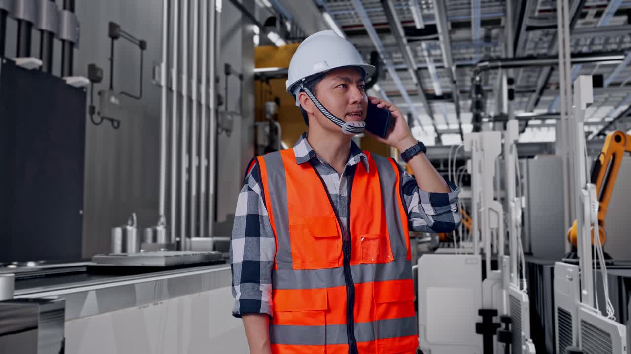 Ingeniero asiático con casco de seguridad hablando por smartphone mientras está de pie, en una línea de producción de baterías automatizada en una fábrica moderna