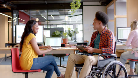 Happy-diverse-female-and-male-colleague-in-wheelchair-with-tablet-talking-at-desk-in-casual-office