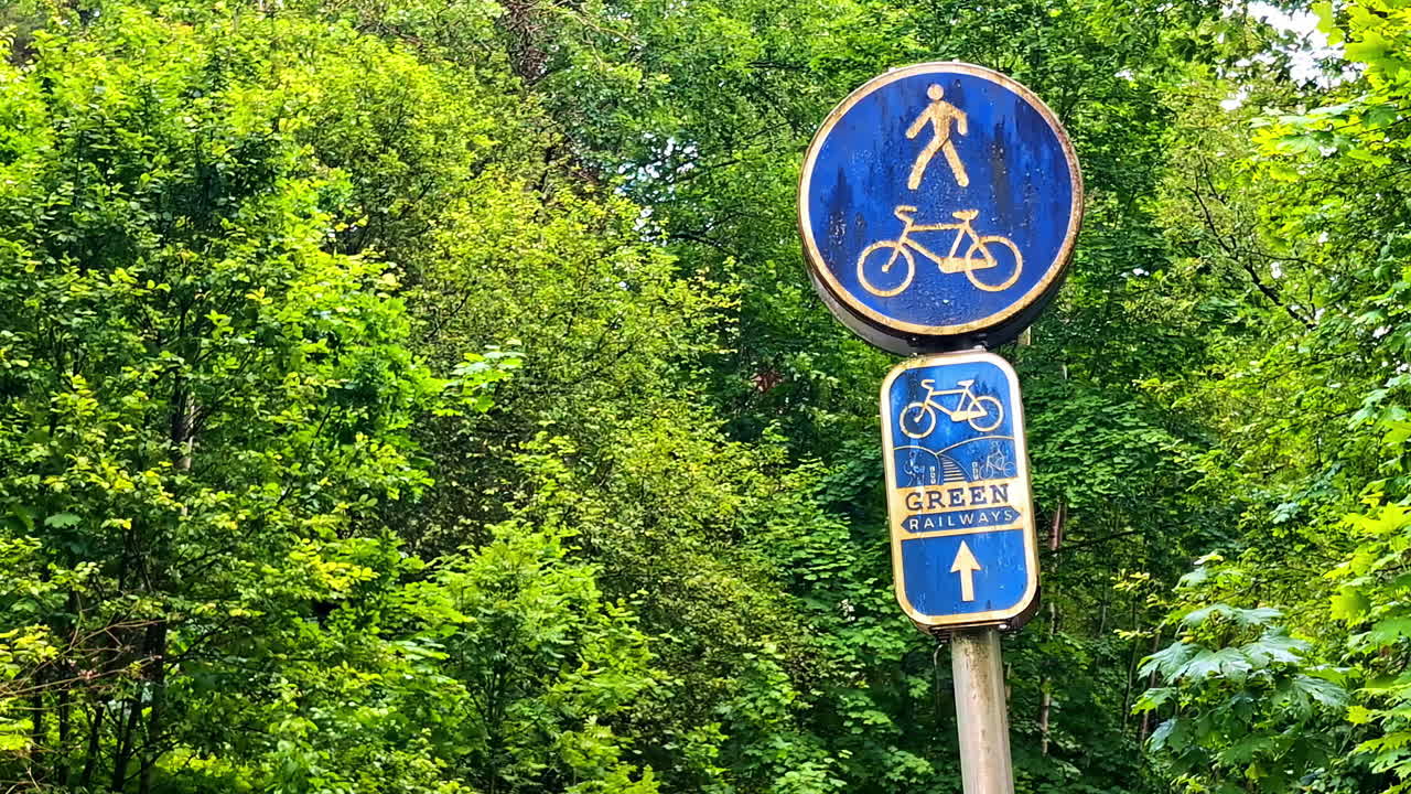 Blue pedestrian and bicycle sign of green railway trail surrounded by forest trees in Latvia
