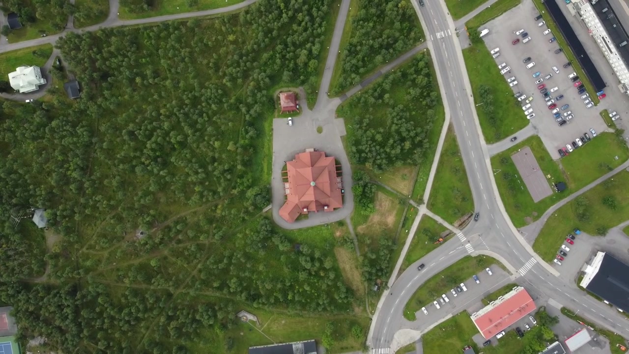 The iconic red wooden Kiruna Church sits atop a hill surrounded by birch trees, winding paths, roads, and parking areas on the edge of town, top down push in