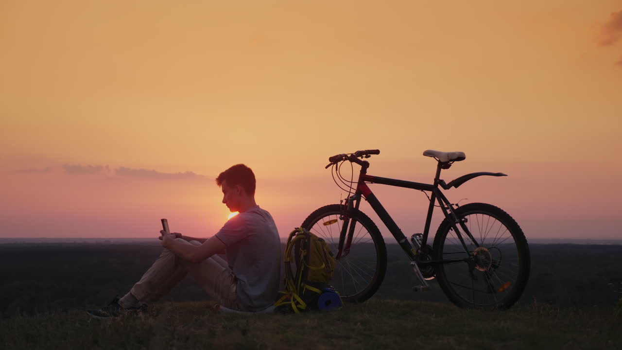 L'adolescente usa uno smartphone, seduto vicino alla sua bicicletta e allo zaino, al tramonto.
