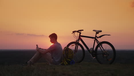 the teenager uses a smartphone sits near his bike and backpack at sunset