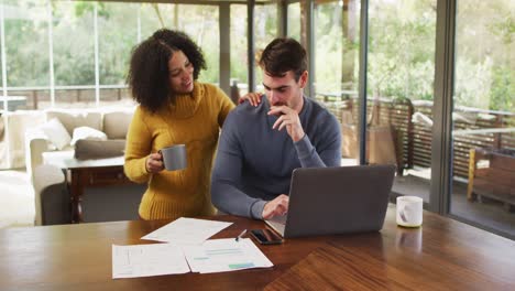 pareja feliz diversa hablando en la sala de estar, usando una computadora portátil, mirando papeles y pagando facturas