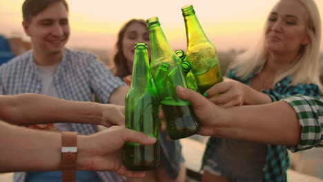 Three-men-and-three-women-in-plaid-shirts-clinks-glasses-and-drinks-beer-from-green-bottels-on-the-party-with-friends-on-the-roof-at-the-sunset.They-are-sitting-together-eat-hot-pizza-after-in-summer-everning.