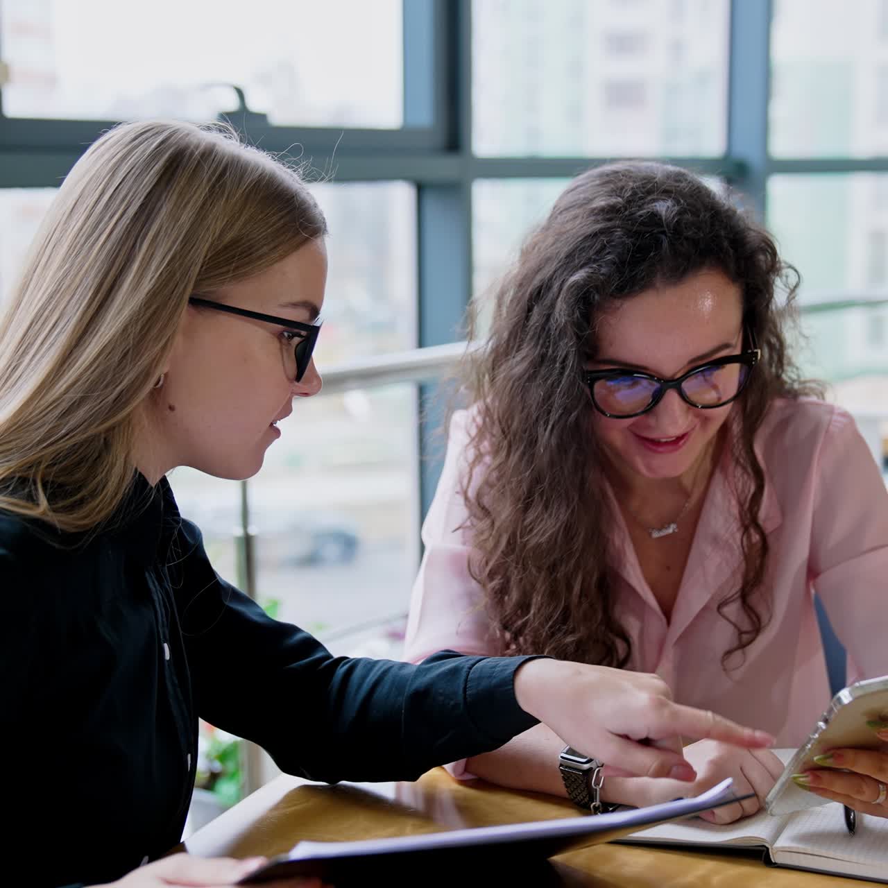 Two young women look at smartphone and discuss something smiling. Female office workers talk about ideas