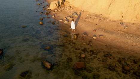 Mujer-Solitaria-Con-Vestido-Blanco-En-La-Playa-Al-Atardecer-Con-Costa-Rocosa-En-Orlowo,-Polonia