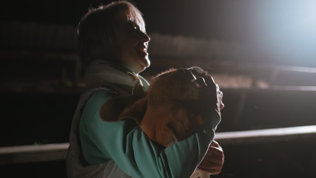 Instructor warmly hugs herd dog playfully under bright spotlight at night creating tender emotional moment showcasing strong bond between human and dog in quiet outdoor setting filled with affection