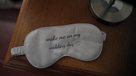 White-sleep-mask-with-"wake-me-on-my-wedding-day"-text,-placed-on-a-wooden-nightstand