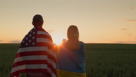 pareja con las banderas de ucrania y los ee.uu. de pie uno al lado del otro y mirar el amanecer sobre un campo de trigo