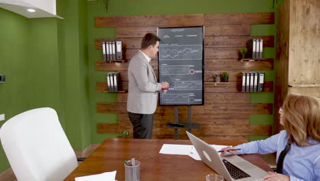 Young-man-holding-a-clipboard-in-front-of-tv-screen-with-charts