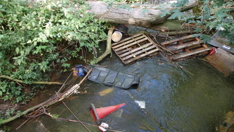 a small stream is cluttered with debris, including wooden pallets, a traffic cone, and branches, all caught under a fallen tree