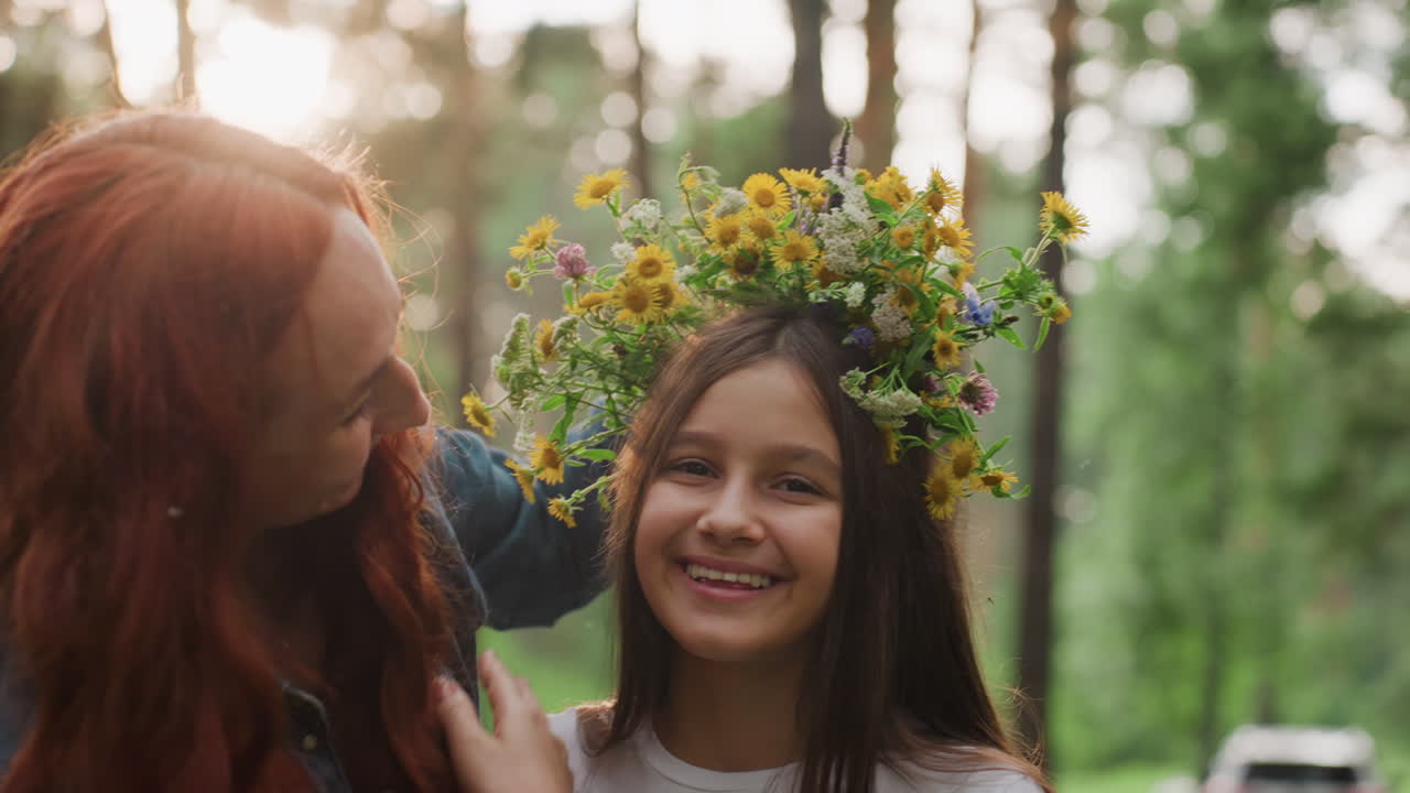 Mother decorates daughter head with wildflowers as child covers eyes with hands, smiling warmly under soft sunlight in peaceful forest, showing joy, love, and emotional family connection outdoors