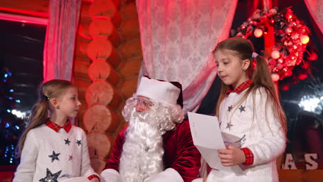 children showing pictures to santa. santa claus with white beard sitting in the room and talking to little girls at christmas. new year night.