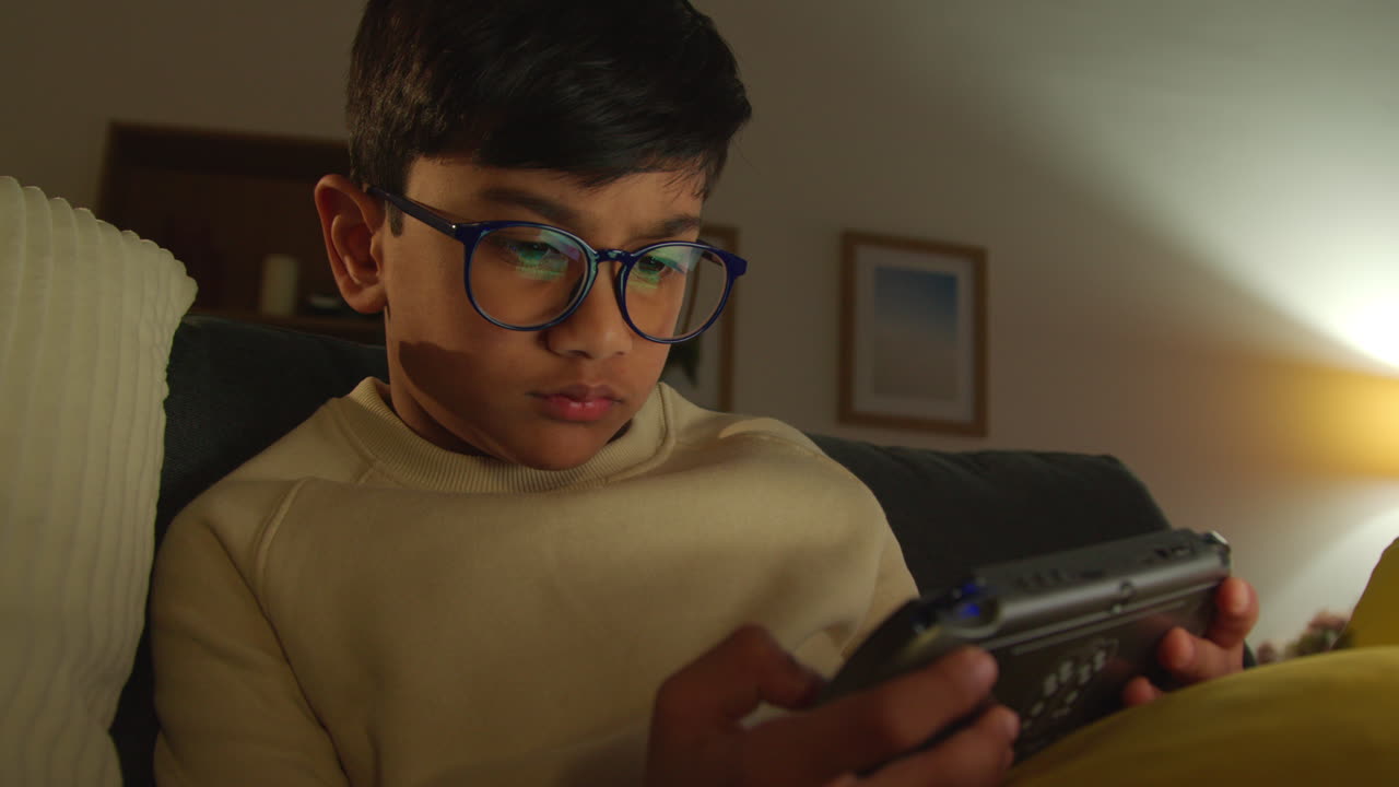 Young Boy Sitting On Sofa At Home Playing Game Or Streaming Onto Handheld Gaming Device At Night 1