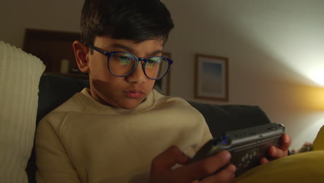 niño sentado en el sofá en casa jugando a un juego o transmitiendo a un dispositivo de juego portátil por la noche 1