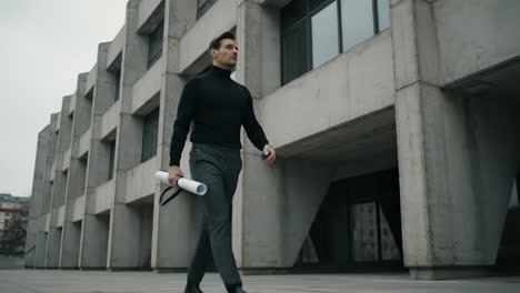 a professional man walks confidently with blueprints in front of a modern concrete building