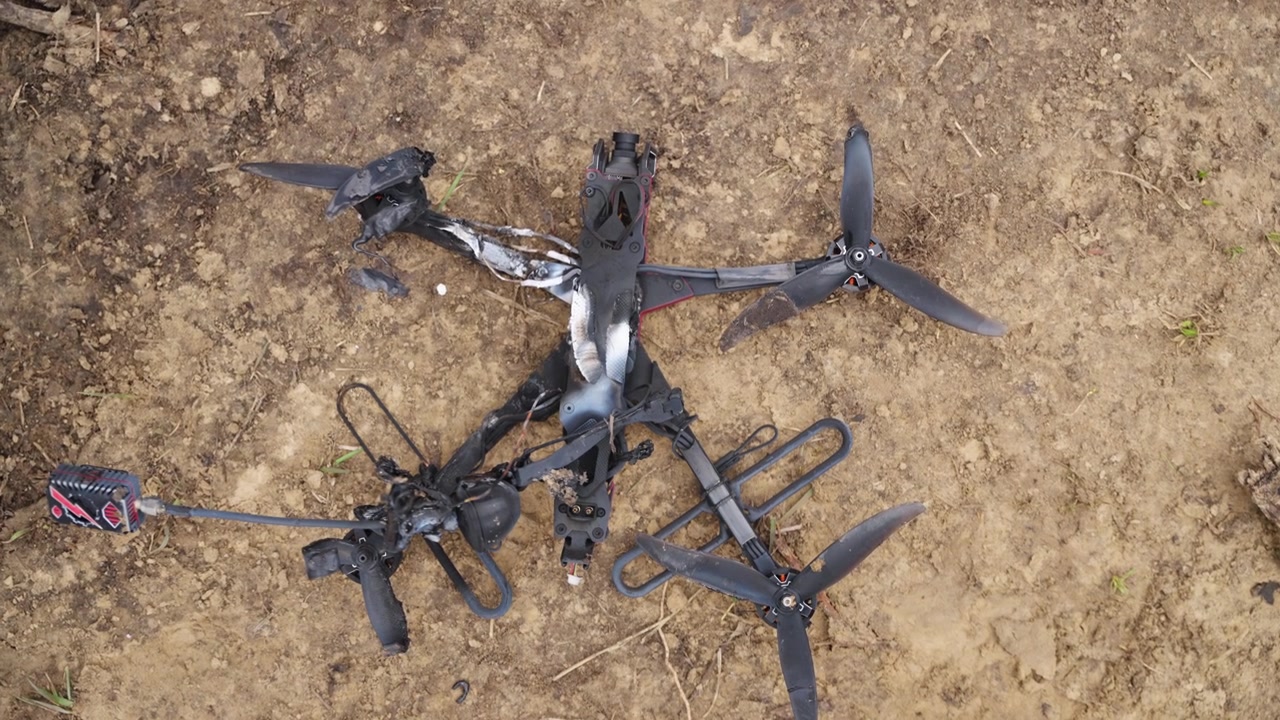 Kerangka drone FPV yang terbakar tergeletak di tanah kotor dengan kerusakan yang terlihat dan bagian yang patah di luar ruangan.