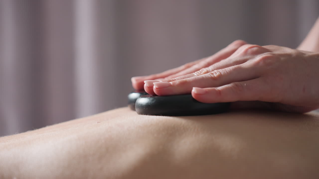 Close view of massage therapist pressing two smooth black stones on client bare back during relaxing therapy session in softly lit spa room with neutral background curtain for calm
