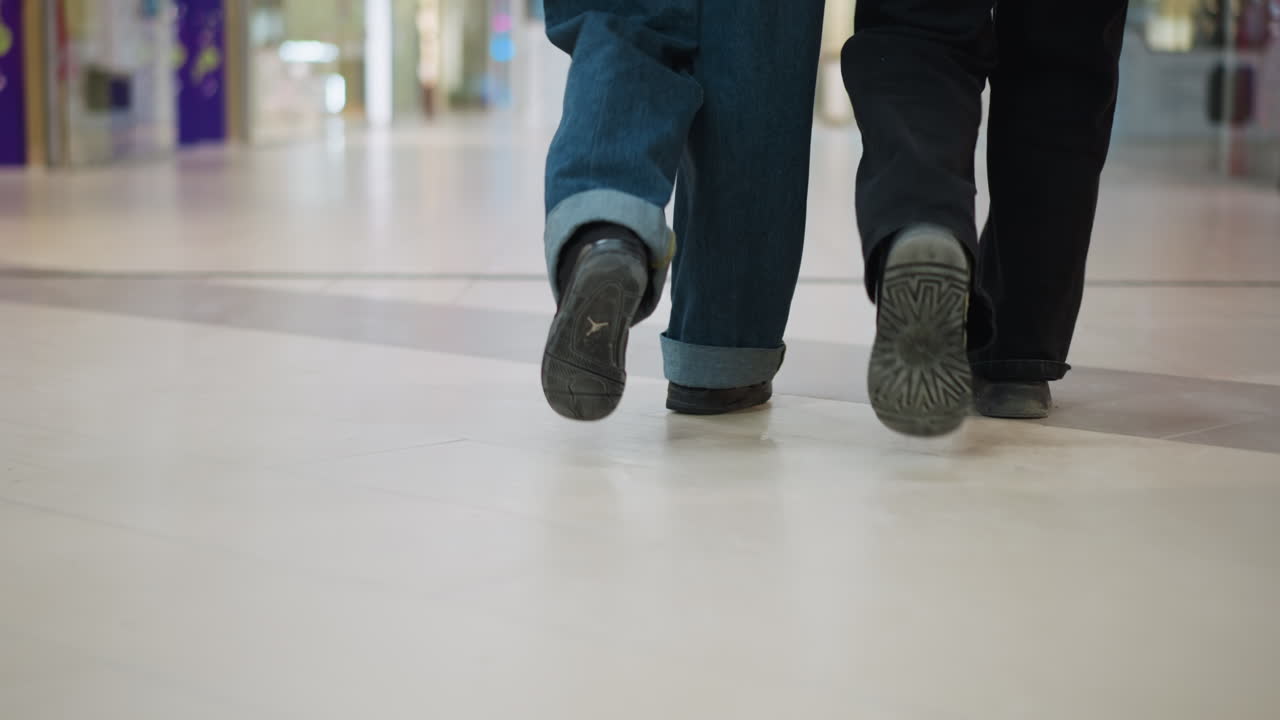 Close up of two adults walking inside shopping mall, casual shoes and wide pants visible on tiled floor, movement captured from behind with blurred shoppers and retail stores in background