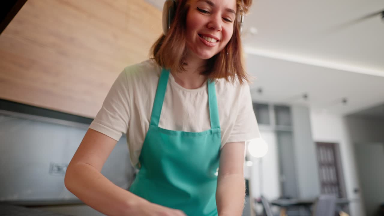 Happy cheerful brunette cleaning lady girl in wireless headphones and a white T-shirt with a blue front washes the stove in the kitchen and dances while having fun in a modern apartment