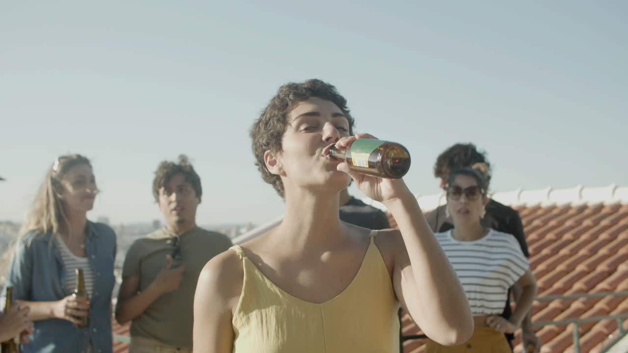 retrato de una mujer caucásica feliz con el pelo corto bebiendo una cerveza y mirando la cámara mientras tiene una fiesta en la azotea con amigos