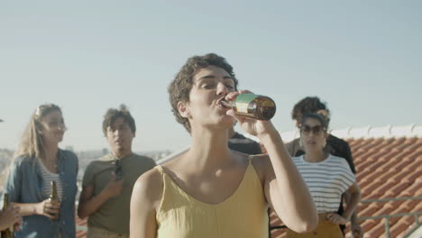 portret van een gelukkige blanke vrouw met kort haar die een biertje drinkt en naar de camera kijkt terwijl ze een feestje op het dak heeft met vrienden